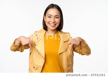 Portrait of happy smiling asian girl, pointing fingers down and showing logo, demonstrating banner, standing in yellow jacket against white background Portrait of happy smiling asian girl, pointing fingers down and showing logo, demonstrating banner, standing in yellow jacket against white background 87032261