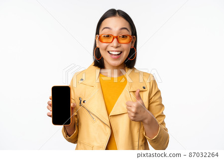 Beautiful korean girl, asian woman in sunglasses, showing smartphone app interface, thumbs up, recommending mobile phone application, white background 87032264