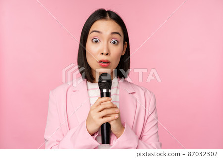 Young asian saleswoman, office lady with suit, holding microphone and looking shocked at camera, talking, giving speech, standing over pink background 87032302