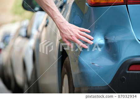 Driver hand examining dented car with damaged fender parked on city street side. Road safety and vehicle insurance concept 87032934