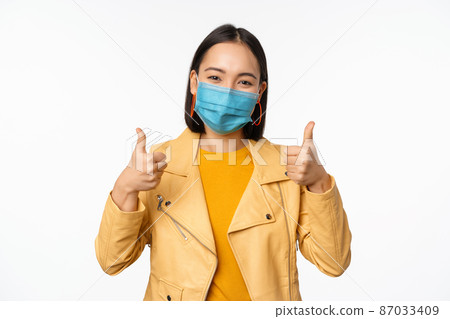 Smiling asian woman in medical face mask showing thumbs up, support wearing protective equipment from covid-19, coronavirus pandemic, white background Smiling asian woman in medical face mask showing thumbs up, support wearing protective equipment from covid-19, coronavirus pandemic, white background 87033409