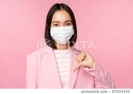 Korean business woman in medical face mask and suit, shows thumbs down, dislike or disapprove gesture, standing over pink background Korean business woman in medical face mask and suit, shows thumbs down, dislike or disapprove gesture, standing over pink background 87033430