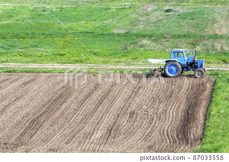 Blue dusty tractor with seedbed cultivator standing at the edge of freshly plowed and cultivated field, soil prepared for sowing. Agriculture, farming and agricultural machinery concept. 87033558