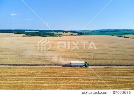 Aerial view of lorry cargo truck driving on dirt road between agricultural wheat fields. Transportation of grain after being harvested by combine harvester during harvesting season Aerial view of lorry cargo truck driving on dirt road between agricultural wheat fields. Transportation of grain after being harvested by combine harvester during harvesting season 87033686