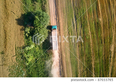 Aerial view of cargo truck driving on dirt road between agricultural wheat fields making lot of dust. Transportation of grain after being harvested by combine harvester during harvesting season 87033765