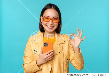 Smiling asian girl with smartphone, shwoing okay, ok sign in approval, standing over blue background 87033863