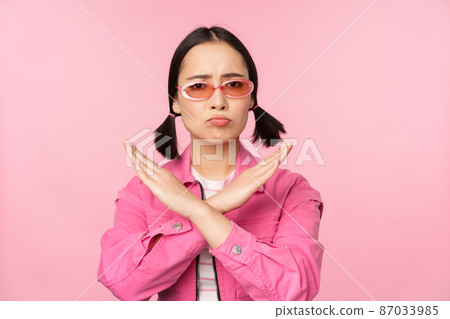 Portrait of korean girl in stylish sunglasses, sulking disappointed, showing stop, rejection gesture, cross sign, standing over pink background 87033985
