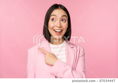 Enthusiastic young asian businesswoman, office employee pointing finger left, looking at banner, advertisement with happy smile, showing advertisement, pink background Enthusiastic young asian businesswoman, office employee pointing finger left, looking at banner, advertisement with happy smile, showing advertisement, pink background 87034447