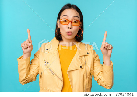 Enthusiastic asian girl in sunglasses, points fingers up, shows banner or logo on top, stands over blue background Enthusiastic asian girl in sunglasses, points fingers up, shows banner or logo on top, stands over blue background 87034841