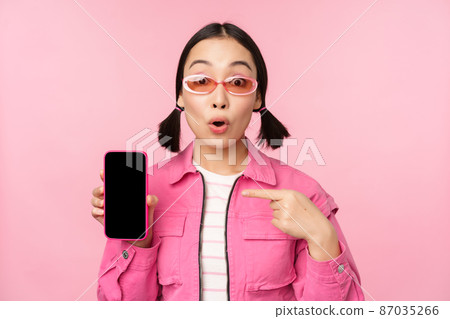 Enthusiastic asian woman in stylish clothes, sunglasses, pointing finger at mobile phone screen, showing smartphone application, standing over pink background Enthusiastic asian woman in stylish clothes, sunglasses, pointing finger at mobile phone screen, showing smartphone application, standing over pink background 87035266