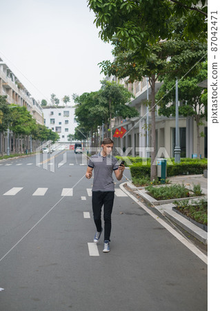 Young caucasian guy walks down the street and makes video call by the phone in wireless headphones. Using smartphone. High quality photo 87042471