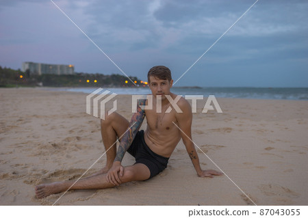 Full-length portrait of handsome caucasian man sitting on the sand on the beach on the sunset, full arm tattoo. High quality photo Full-length portrait of handsome caucasian man sitting on the sand on the beach on the sunset, full arm tattoo. High quality photo 87043055