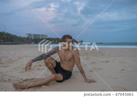 Full-length portrait of handsome caucasian man sitting on the sand on the beach on the sunset, full arm tattoo. High quality photo 87043061