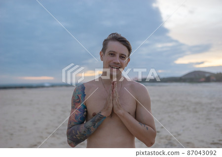 portrait of handsome fit bare-chested young man standing on the beach, guy with tattoo on sunset. Smiling and holding hands near his breast.  87043092