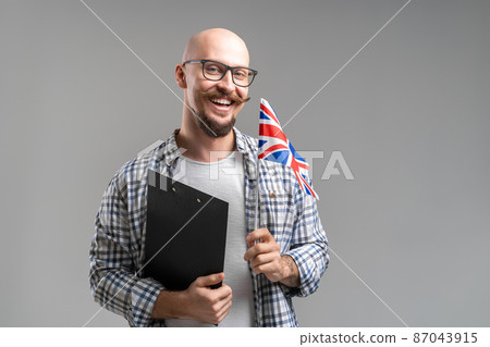 Smiling student holds an United Kingdom flag and clipboard while isolated on gray background Smiling student holds an United Kingdom flag and clipboard while isolated on gray background 87043915