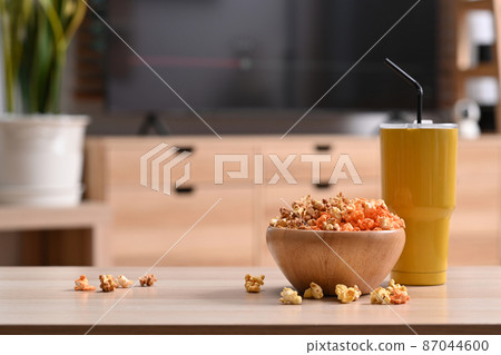 A wooden bowl with fresh popcorn and glass on the background of a home interior with a TV. 87044600