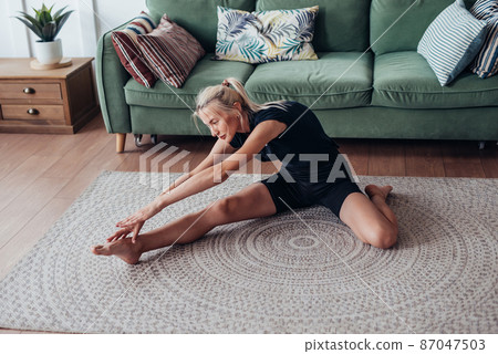 Woman working out at home, doing stretching bends forward while sitting on floor 87047503