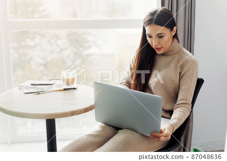 Beautiful young woman working on laptop computer while sitting at the living room, drinking coffee. Beautiful young woman working on laptop computer while sitting at the living room, drinking coffee. 87048636