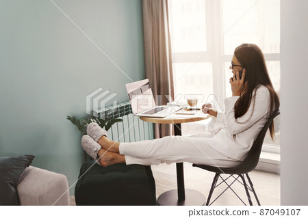 Beautiful woman talking on phone while  working on laptop computer sitting at home managing her business via home office during Coronavirus or Covid-19 quarantine 87049107