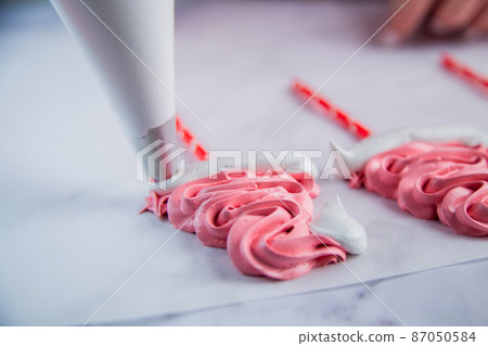 On light parchment there are pink meringue caps on sticks, the cook makes a pompom and a frill from a pastry bag of white meringue. Photography in a light key On light parchment there are pink meringue caps on sticks, the cook makes a pompom and a frill from a pastry bag of white meringue. Photography in a light key 87050584