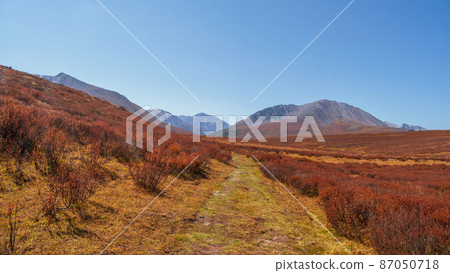 Dirt road through the autumn mountain plateau. Rough dirt road leading through orange autumn fields into mountains beyond under blue sky. Panoramic view. 87050718