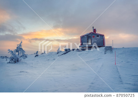 Cabin in winter dawn. Snow-covered staircase leading through snowdrifts to a lonely house on a hilltop in the cool evening. Cabin in winter dawn. Snow-covered staircase leading through snowdrifts to a lonely house on a hilltop in the cool evening. 87050725