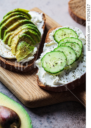 Toasts with cream cheese, avocado, cucumber and rye bread on wooden board, dark background. 87052617