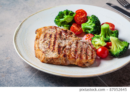Grilled pork steak with broccoli and tomatoes on plate, dark background. 87052645
