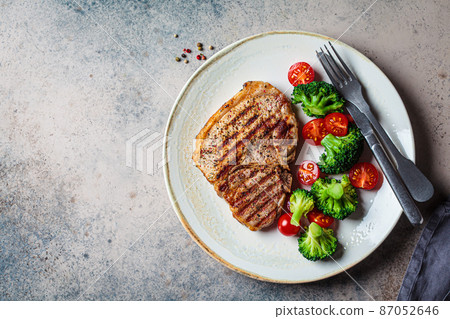 Grilled pork steak with broccoli and tomatoes on plate, dark background. 87052646