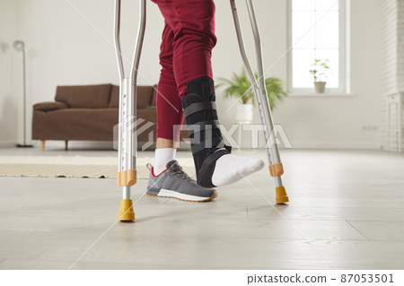 Injury and rehabilitation. Close up of legs of man who uses crutches to walk around room as he has leg injury. Uunknown man with ankle injury wears special bandage on his leg and uses metal crutches. Injury and rehabilitation. Close up of legs of man who uses crutches to walk around room as he has leg injury. Uunknown man with ankle injury wears special bandage on his leg and uses metal crutches. 87053501