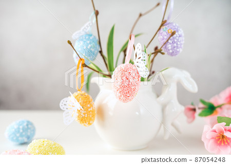 Home interior with easter decor. Vase with willow tree branches with Easter eggs and bunny on white table and background with copy space Home interior with easter decor. Vase with willow tree branches with Easter eggs and bunny on white table and background with copy space 87054248