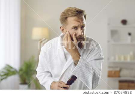 Portrait of man who examines his face in morning checking condition of skin and presence of wrinkles. Handsome man in bathrobe with hairbrush in his hand looks at camera. Concept of male beauty. 87054858