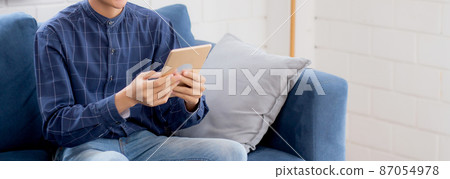 Young attractive asian man resting using browsing tablet computer on sofa at home. Young attractive asian man resting using browsing tablet computer on sofa at home. 87054978