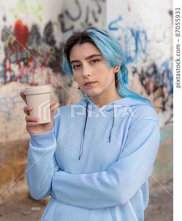 Blue haired Teenage girl in light blue oversize hoodie staying near graffiti wall with coffee to go Blue haired Teenage girl in light blue oversize hoodie staying near graffiti wall with coffee to go 87055931