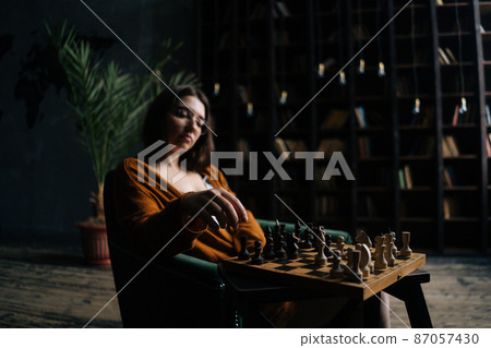 Portrait of cofident young business woman wearing elegant eyeglasses making chess move sitting in armchair in dark library room. Pretty intelligent lady playing logical board game alone at home. 87057430