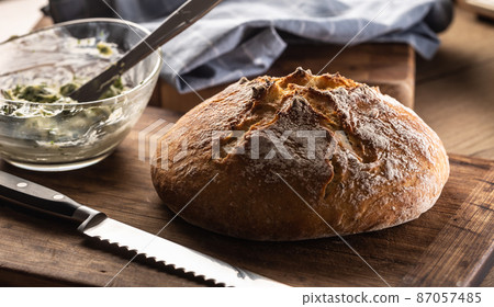 Crusty bread loaf next to a knife and butter mixed with herbs in a bowl Crusty bread loaf next to a knife and butter mixed with herbs in a bowl 87057485