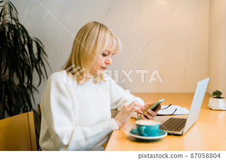 Mature adult woman sitting in cafe with coffee mug and working online on laptop computer. Businesswoman texting messages at mobile phone in coworking space in roasters coffee shop 87058804