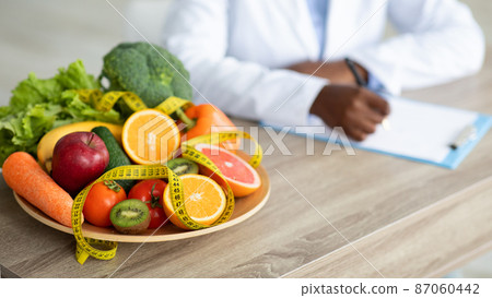 Black nutritionist writing diet plan, sitting at workplace in clinic, focus on plate with fruits and vegetables, cropped Black nutritionist writing diet plan, sitting at workplace in clinic, focus on plate with fruits and vegetables, cropped 87060442
