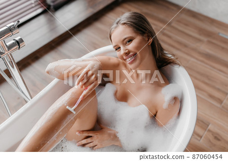 Spa treatment, hair removal. Happy lady shaving her legs in bubble bath, using razor, removing unwanted body hair Spa treatment, hair removal. Happy lady shaving her legs in bubble bath, using razor, removing unwanted body hair 87060454