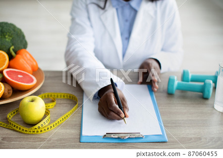 Cropped view of black female dietitian planning meal for client, writing in clipboard, sitting at desk at clinic Cropped view of black female dietitian planning meal for client, writing in clipboard, sitting at desk at clinic 87060455