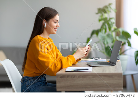 Happy young woman in earphones sitting at desk with laptop, using cell phone at home, blank space Happy young woman in earphones sitting at desk with laptop, using cell phone at home, blank space 87060478