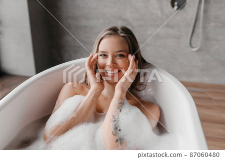 Skin care and spa treatments at home. Happy relaxed lady in bathtub with foam, touching her smooth face skin and smiling Skin care and spa treatments at home. Happy relaxed lady in bathtub with foam, touching her smooth face skin and smiling 87060480