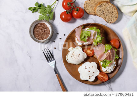Easter breakfast or brunch. Delicious breakfast or snack - poached egg and cream cheese toast whole grain rye bread, prosciutto, arugula on a marble tabletop. Top view flat lay. Copy space. 87060481