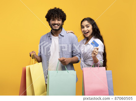 Consumerism concept. Excited indian couple with credit card and shopping bags, buying remotely, yellow background 87060505