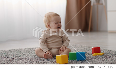 Cute little upset toddler sitting alone on floor with colorful plastic cubes and crying, looking aside for mommy 87060549