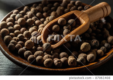 Allspice jamaica pepper in a wooden scoop over allspice grains on a wooden plate. 87061408