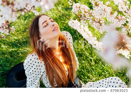 Young brunette woman in dress with closed eyes sitting near a pink blooming tree and enjoying the sunshine. Generation Z girl enjoy spring mood during a sunny day. Relax on the nature. 87063025