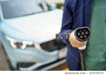 Asian man preparing to charge his electric car or EV at charging station, man holding a Type 2 BEV charging connector. Electric Vehicle or EV car and zero emission concept. 87070554