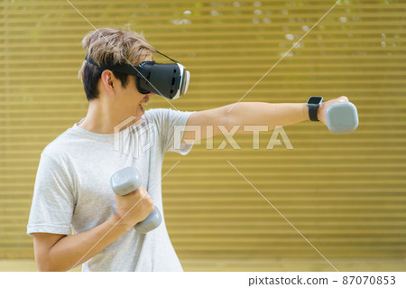 Active Asian sportsman doing a weight training exercise by using dumbbell and virtual reality - VR goggles platform for online training. The innovation of virtual reality or metaverse platform concept 87070853
