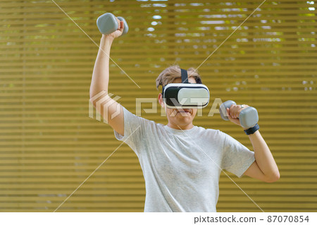 Active Asian sportsman doing a weight training exercise by using dumbbell and virtual reality - VR goggles platform for online training. The innovation of virtual reality or metaverse platform concept 87070854